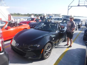 A group of people with their Mazda MX-5 Miata convertibles parked on a ferry with waterfront views.