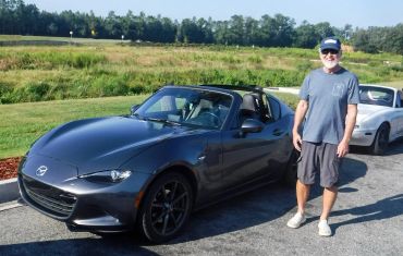 Smiling man standing beside a gray Mazda convertible on a sunny day.