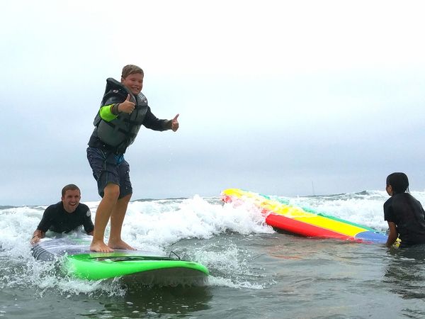 Joey throwing up 2 thumbs up as he rides in on the big green surfboard with Alec on the tail.