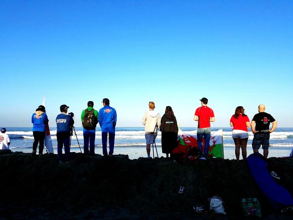 Group of surfers and photographers lined up across the sand watching the action in the ocean.