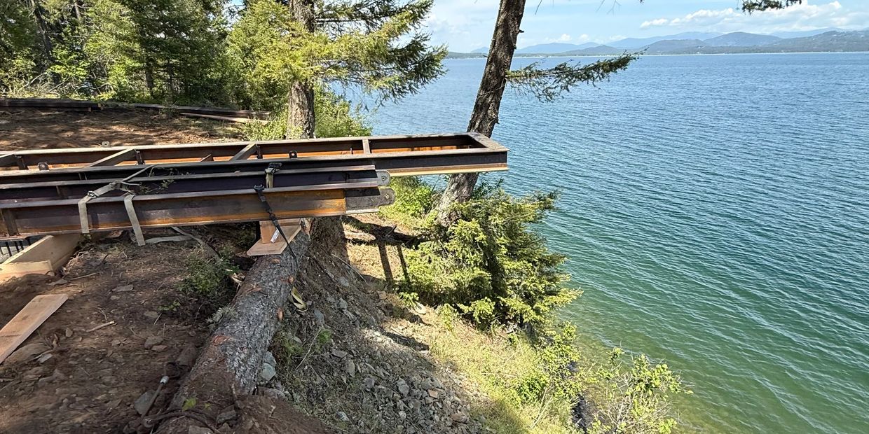 Metal framework extends over a steep lakeside slope surrounded by trees.