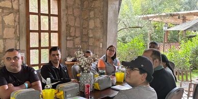 Group of six people sitting around a table indoors near a garden.