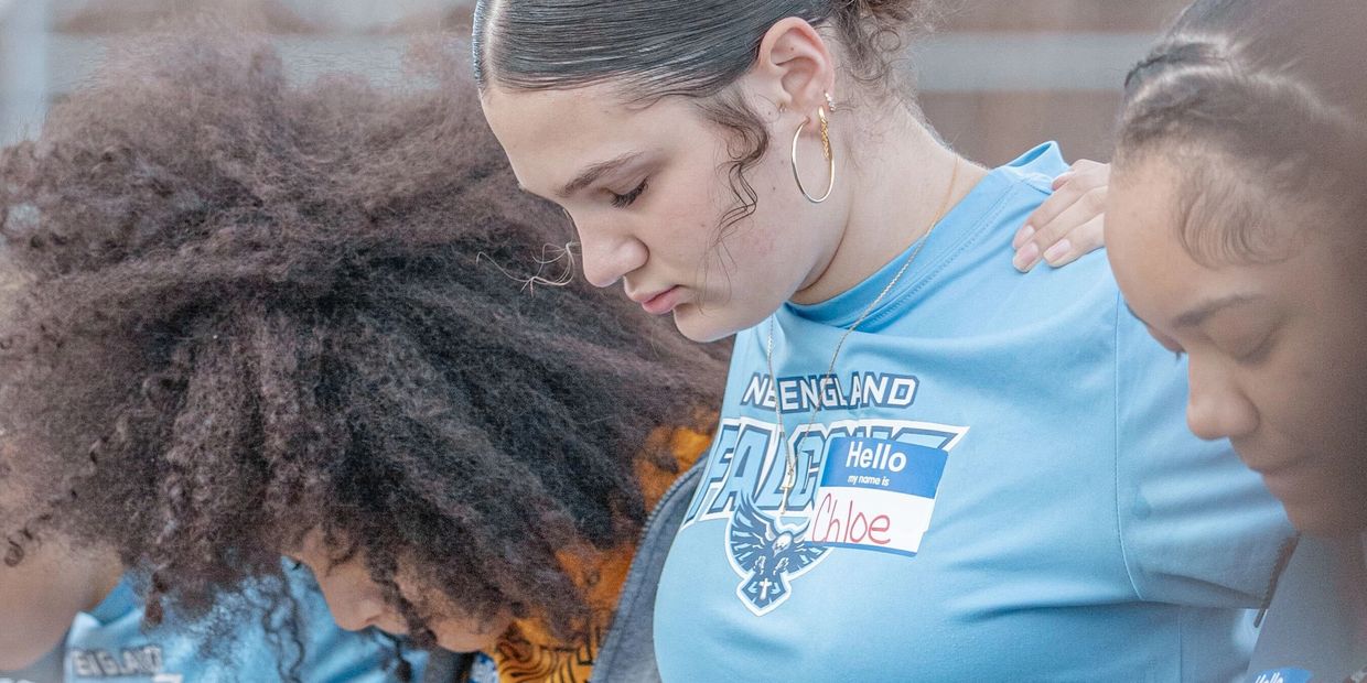 Three young women in blue shirts standing close together with heads bowed.