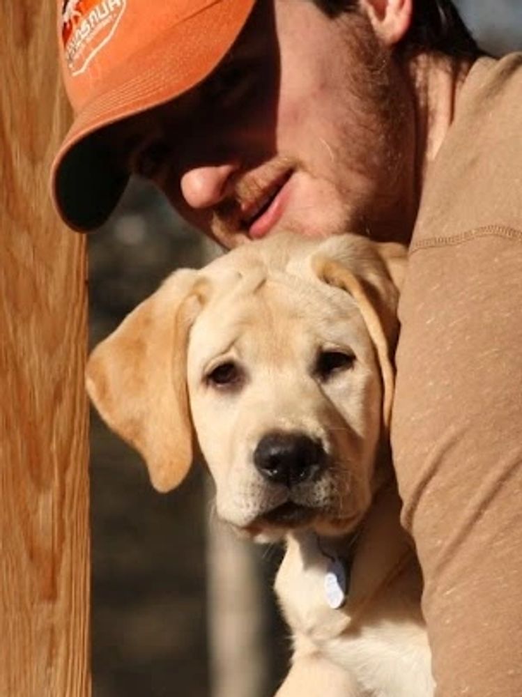 A man in a cap embraces a Labrador puppy on a wooden porch.