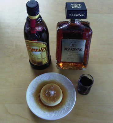 Two bottles of liqueurs and a plate with caramel flan and a shot glass on a wooden table.