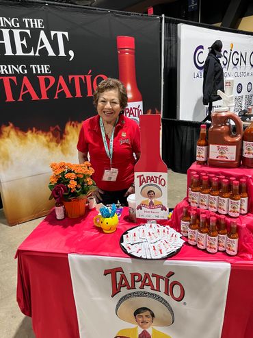 Smiling woman at a vibrant Tapatío hot sauce booth with various sauces and promotional items.