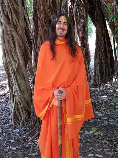 A smiling man in traditional orange attire stands with a staff in a forested area.