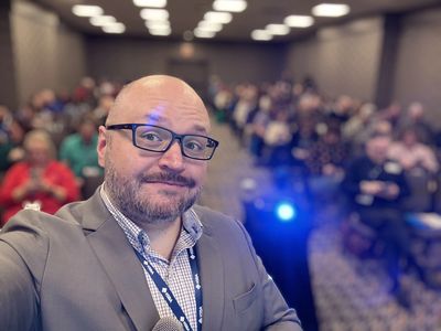 Man in suit taking selfie with audience in background.