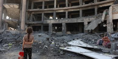 A child stands amidst rubble in front of a destroyed building.