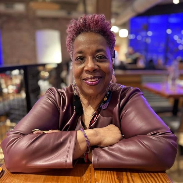 Smiling woman with purple hair wearing a maroon jacket.