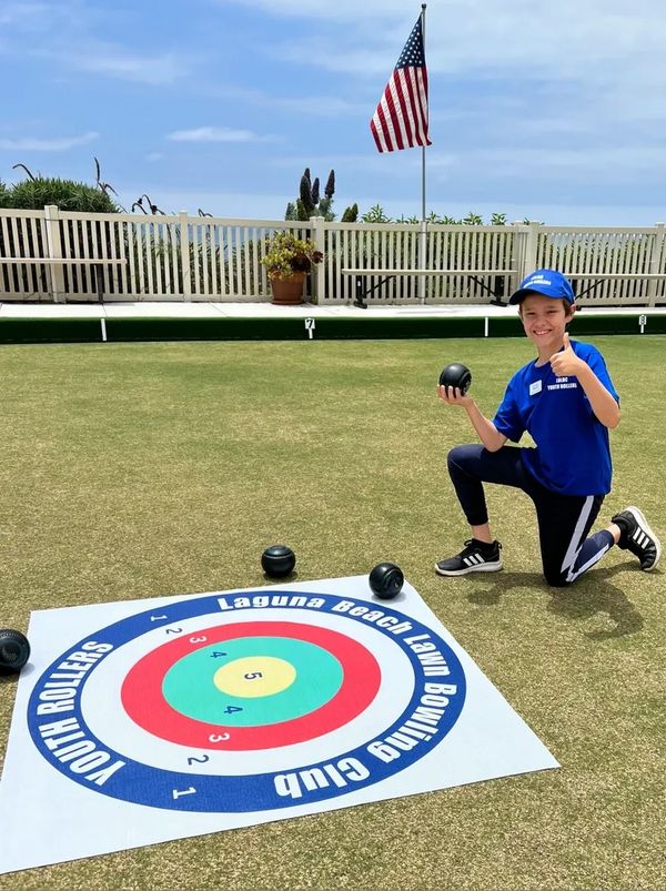 A boy in blue sportswear kneels on a lawn bowling field, holding a ball and giving a thumbs-up.