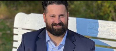 A man with a beard in a navy blazer sitting on a white bench outdoors.