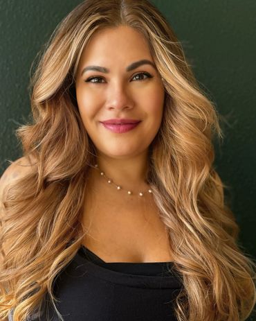 Smiling woman with long wavy blonde hair and a pearl necklace against a dark green background.