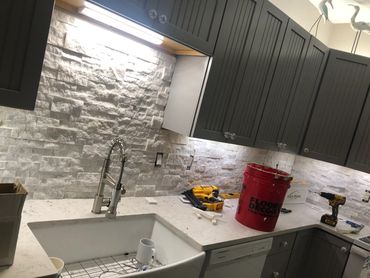 Kitchen countertop with tools and a red bucket under gray cabinets and stone backsplash.