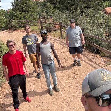 A group of five men posing on a dirt trail with trees and a wooden fence.