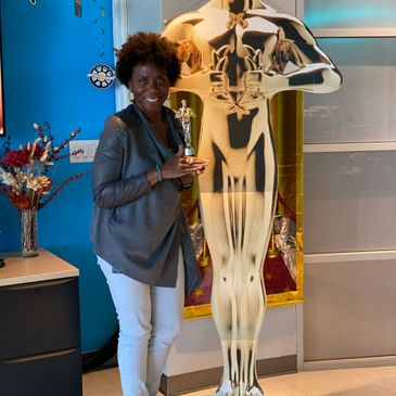 Trish in an office setting, standing near an Oscar statuette standee, holding a mini-replica statuet