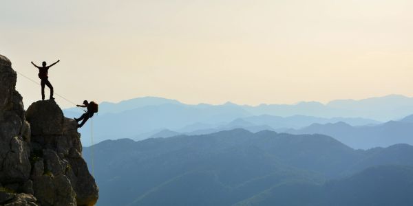 Two climbers on a rocky cliff with mountains in the background.