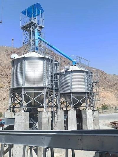 Two large metal grain silos with blue conveyor system in a mountainous area.