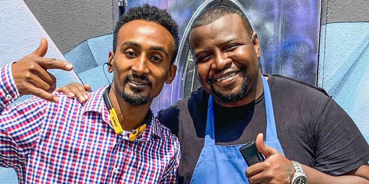 Two men smiling and posing together, one in a checkered shirt and the other in a blue apron.