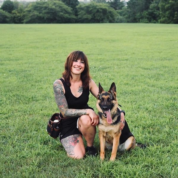 Smiling woman with tattoos kneeling beside a happy German Shepherd in a green field.