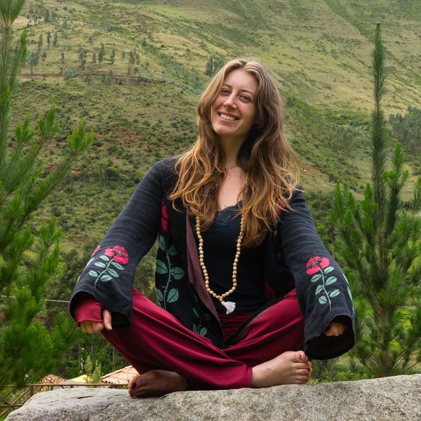 A woman meditates outdoors on a rock, smiling and surrounded by greenery.