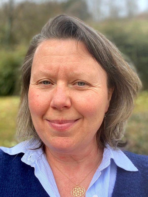 A middle-aged woman smiling outdoors with a blue sweater and necklace.