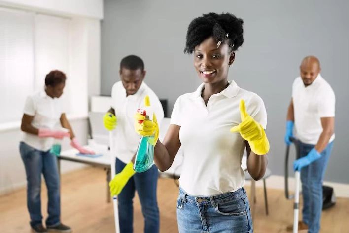 A smiling woman in yellow gloves holding a spray bottle, with three people cleaning in the background.