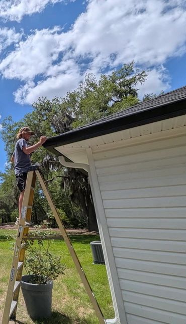 Man standing on ladder fixing or inspecting roof gutter outdoors.