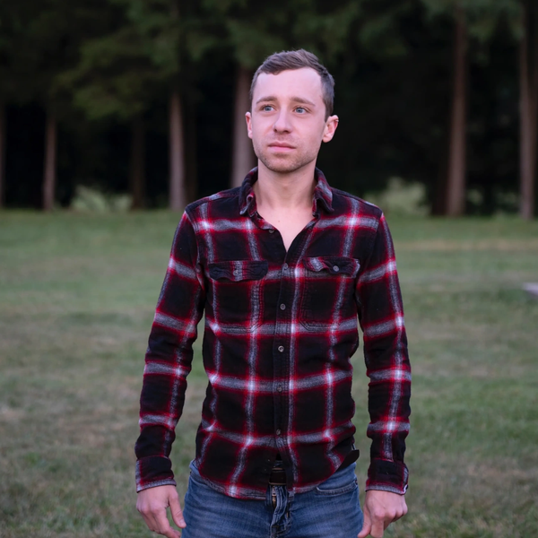 Man wearing a red and black plaid shirt standing outdoors on grass.