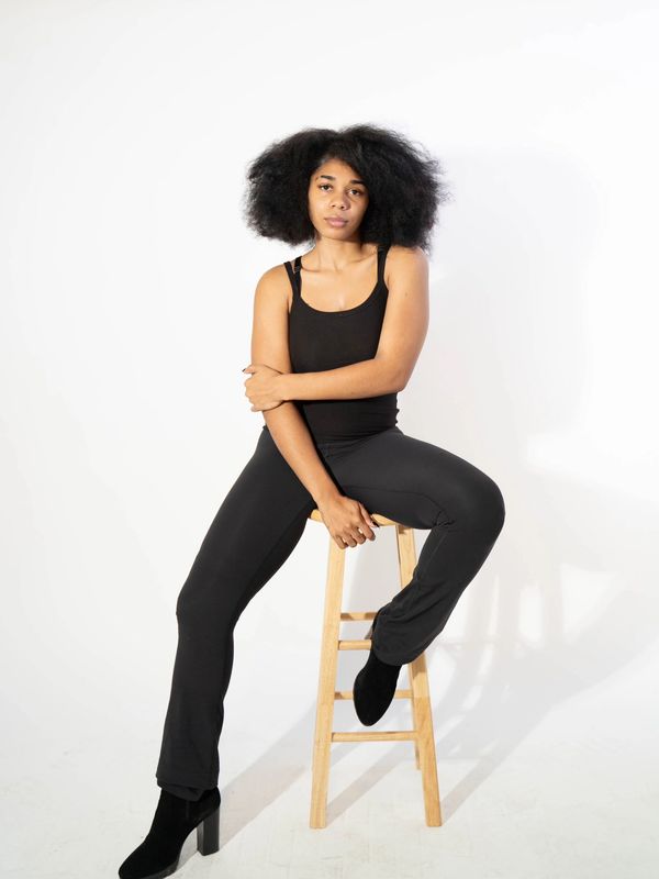 Woman in black outfit sitting on a wooden stool against a white background.