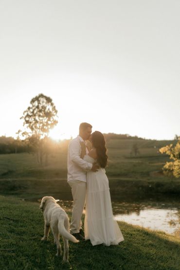 Couple kissing at sunset by a pond with a dog nearby.