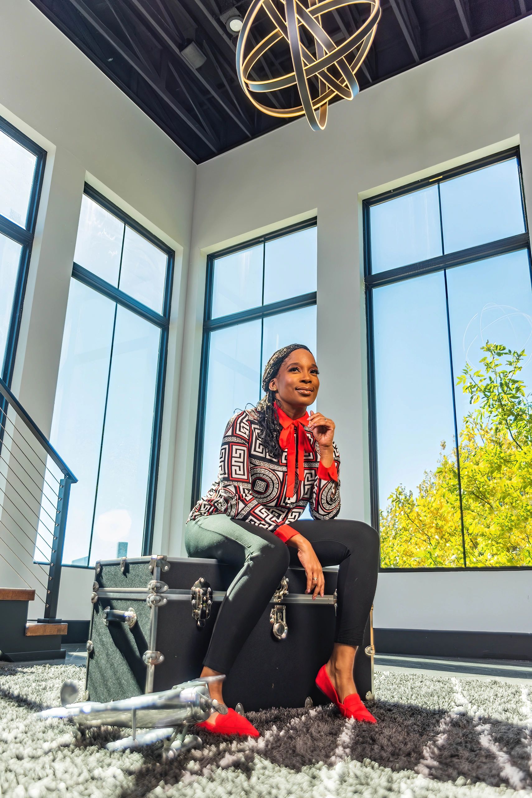 Woman in stylish outfit sitting on a black trunk in a bright room with large windows.