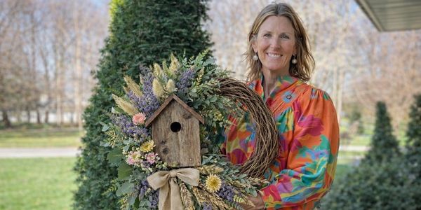 Woman holding a floral wreath with a birdhouse centerpiece outdoors.