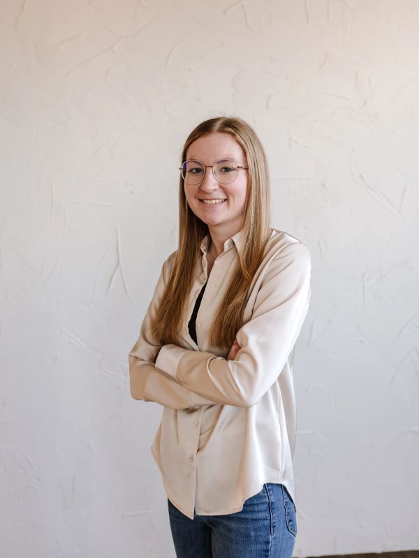 Young woman with glasses and long blonde hair smiling with arms crossed.