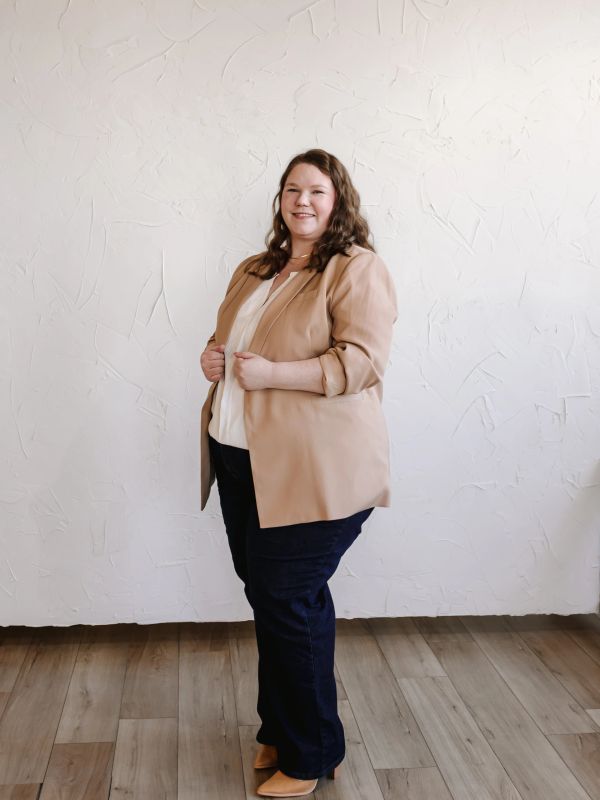 Confident woman in beige blazer and dark jeans against textured white wall.