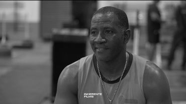 Black and white photo of a man smiling, wearing a sleeveless shirt and chain necklace.