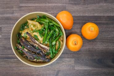 Bowl with grilled fish, pasta, asparagus, and green onions, next to three oranges on a wooden surface.