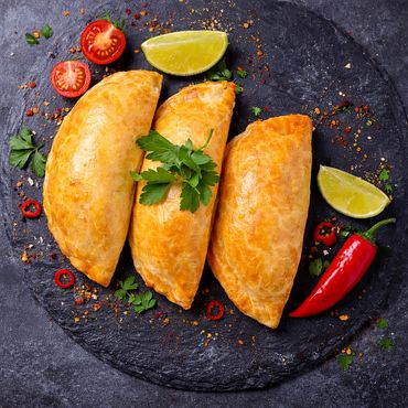 Three golden empanadas garnished with herbs, lime wedges, and chili peppers on a dark slate plate.