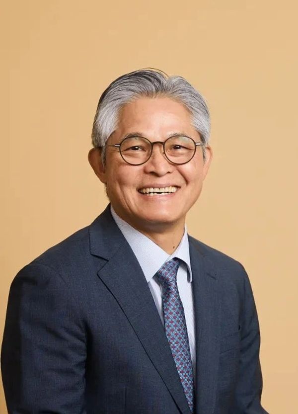 Smiling elderly man in a suit and glasses against a beige background.
