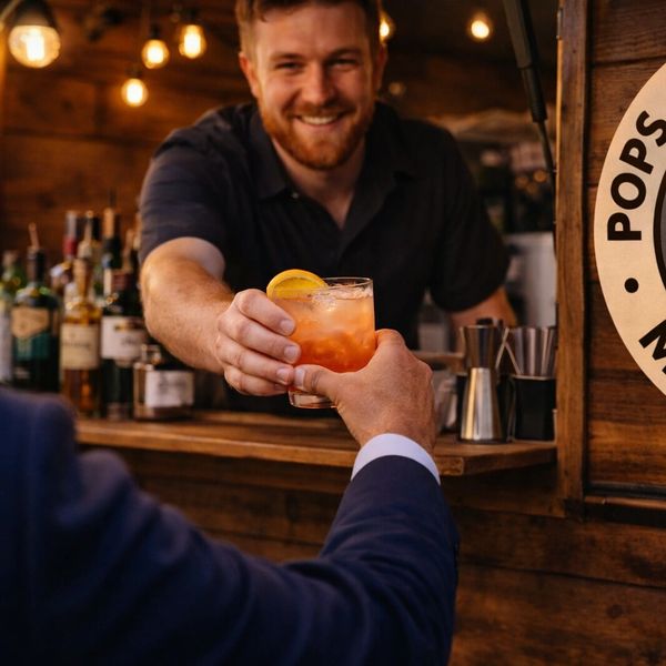 Bartender smiling while handing a cocktail to a customer at a wooden bar.