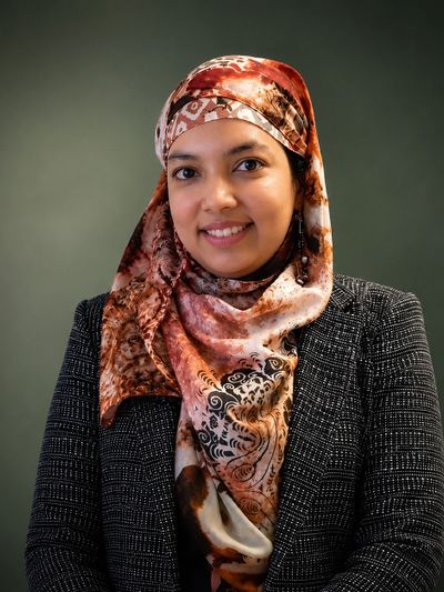 Smiling woman wearing a patterned hijab and checkered blazer against a plain background.
