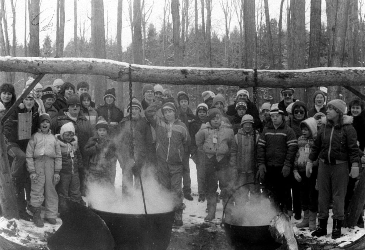 Group of children and adults gathered around steaming kettles in a snowy forest.