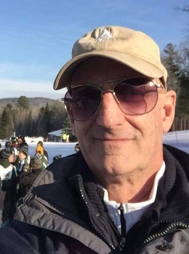 William "Brett" Mayfield. A senior white man with sunglasses and ball cap smiles in the snow.
