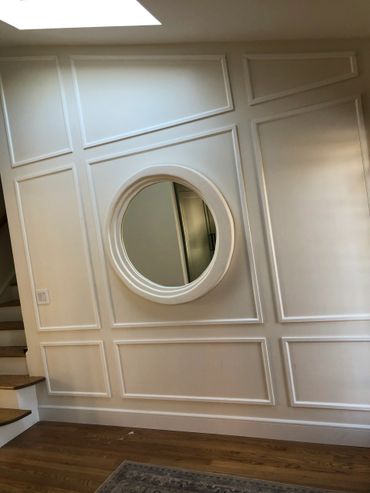 Round mirror on a white paneled wall near wooden stairs.