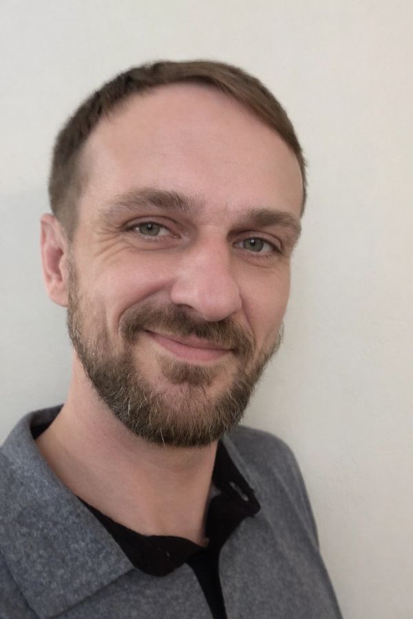 Smiling man with a beard wearing a gray shirt against a plain background.