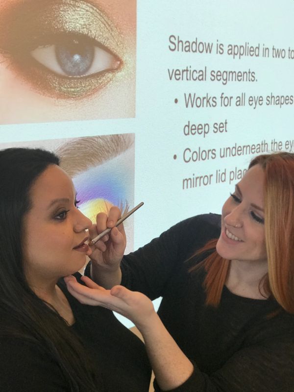 Instructor applying lip gloss with a lipbrush onto a model in front of a classroom