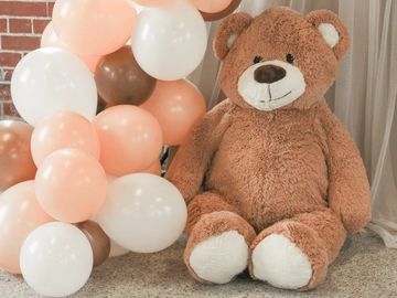 Large teddy bear sitting next to pastel and brown balloons.
