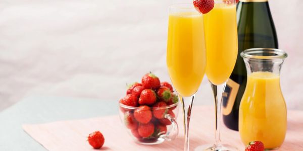 Two mimosa glasses garnished with strawberries beside a bottle and a cup of fresh strawberries.