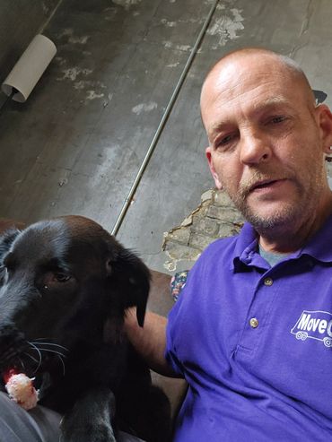 Man in purple shirt with a black dog holding a toy indoors.