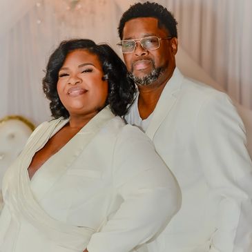 A stylish couple dressed in elegant white suits pose confidently indoors.